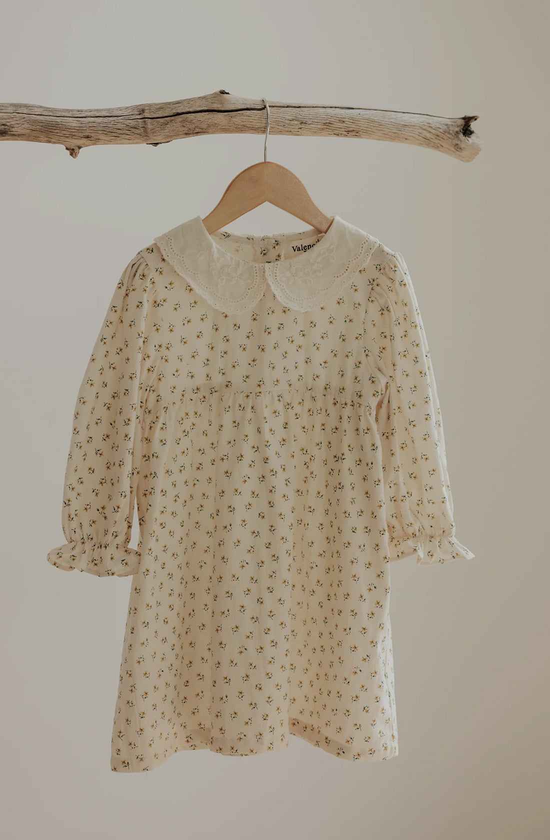 The VALENCIA BYRON BAY's The Farmer's Daughter’s Dress is a breathable mid-weight, cream-colored child's dress adorned with a delicate floral print and elegantly hung on a rustic wooden branch. This timeless classic features a lace-trimmed Peter Pan collar and long sleeves with chic ruffled cuffs.