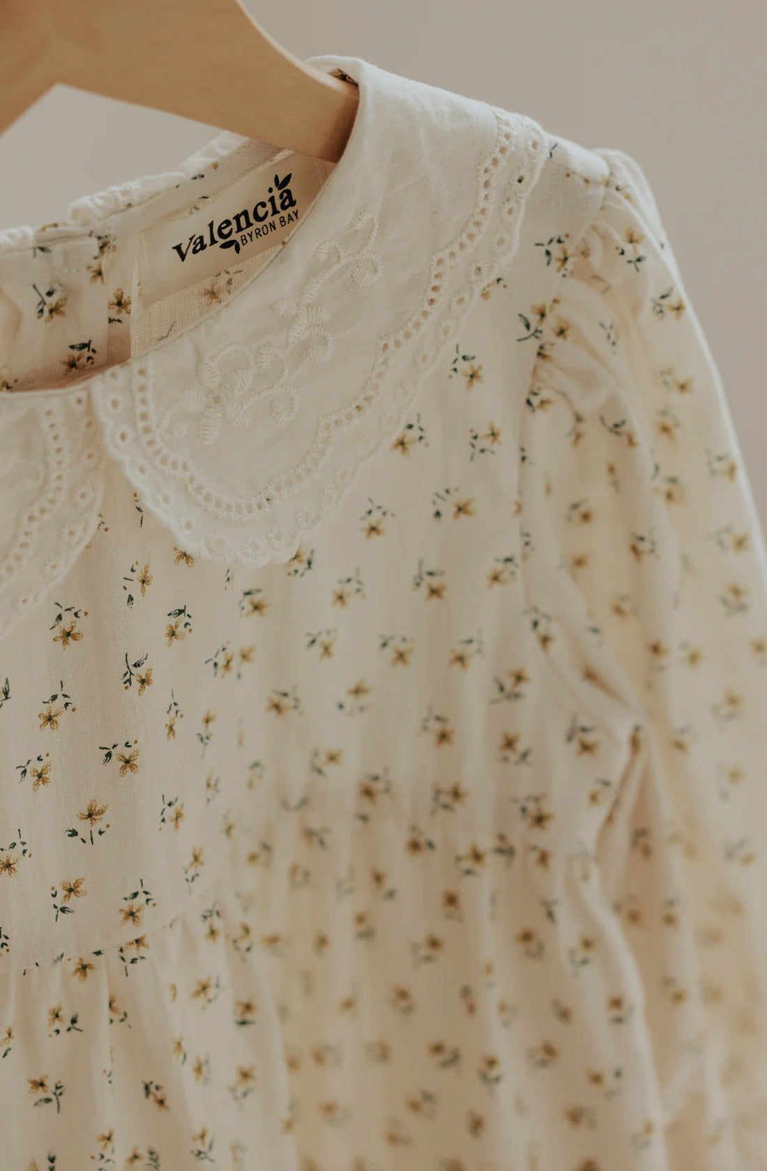 A close-up of a cream-colored children's dress, known as The Farmer's Daughter's Dress, with a delicate floral pattern hangs on a wooden hanger. It features a white, lace-trimmed Peter Pan collar and is labeled "VALENCIA BYRON BAY" on the tag inside the neckline. The breathable mid-weight fabric gathers slightly at the chest.