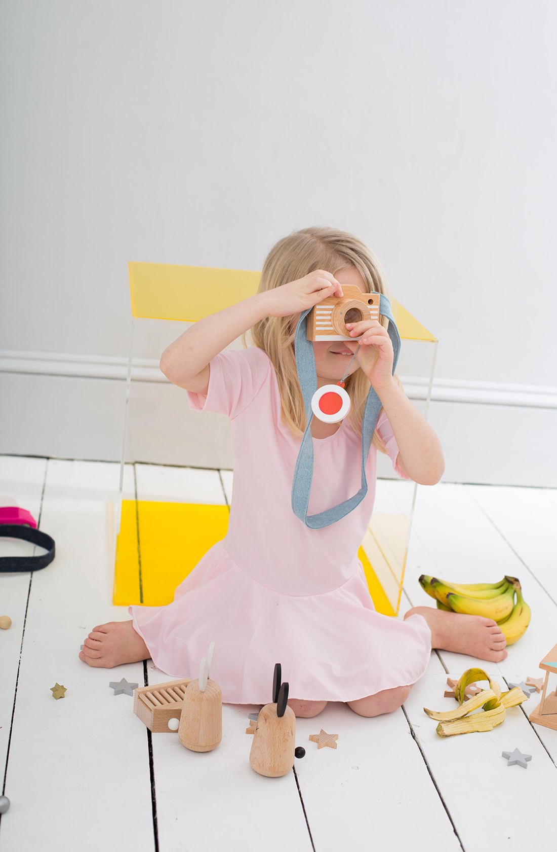 A child with blonde hair, wearing a pink dress, holds the KIKO & GG Kaleidoscope Play Camera in Pink up to their face like a budding toy photographer. The child sits on a white floor scattered with various toys, including wooden animals, a yellow box, and bananas.