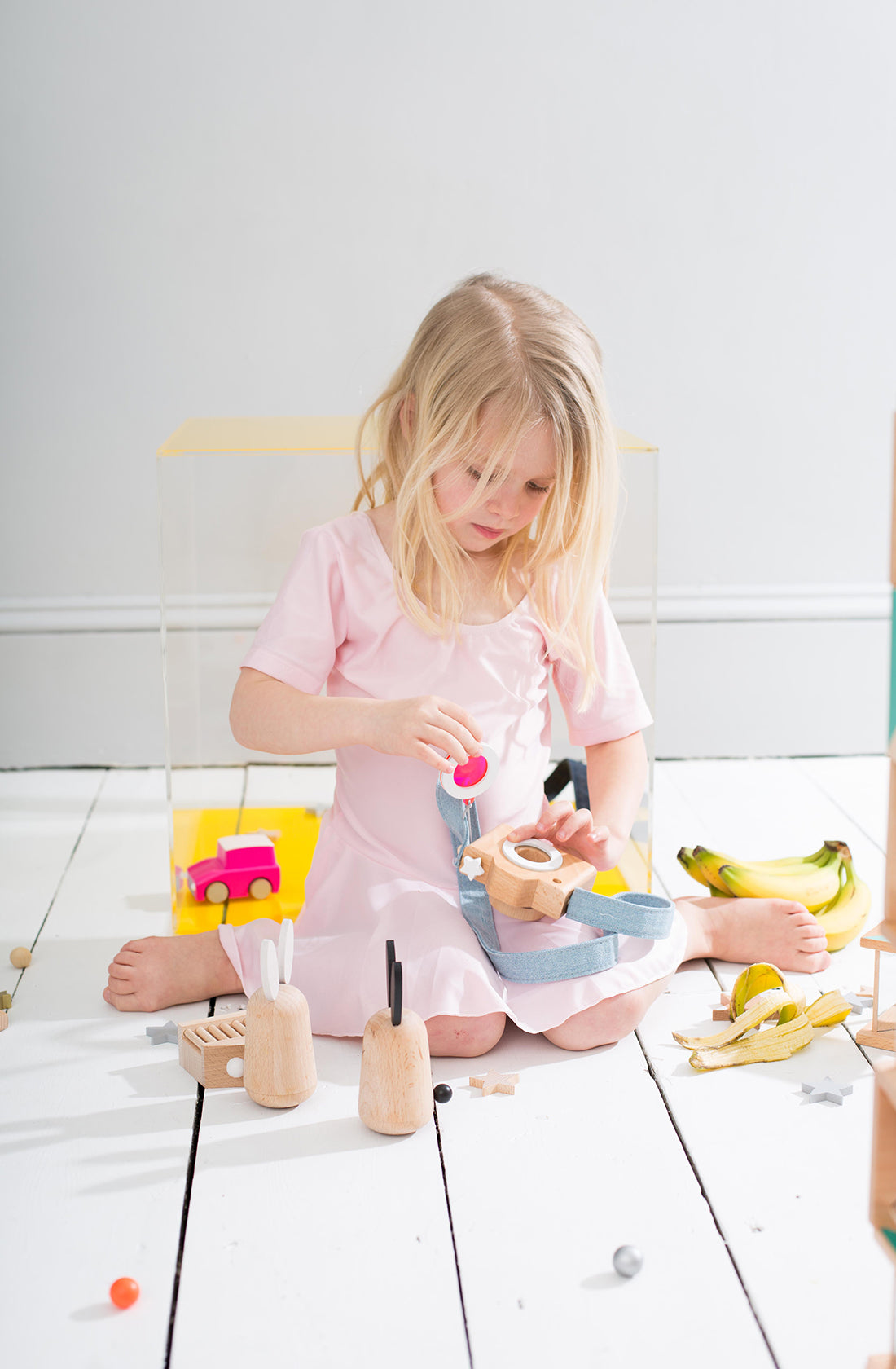 A young child with long blonde hair, wearing a pink dress, sits on a white wooden floor. They are engrossed in playing with a toy tea set. Scattered around them are toy food items, including a banana, and various wooden toys. A pink toy car is visible in the background, captured by a keen toy photographer through their KIKO & GG Kaleidoscope Play Camera Pink.