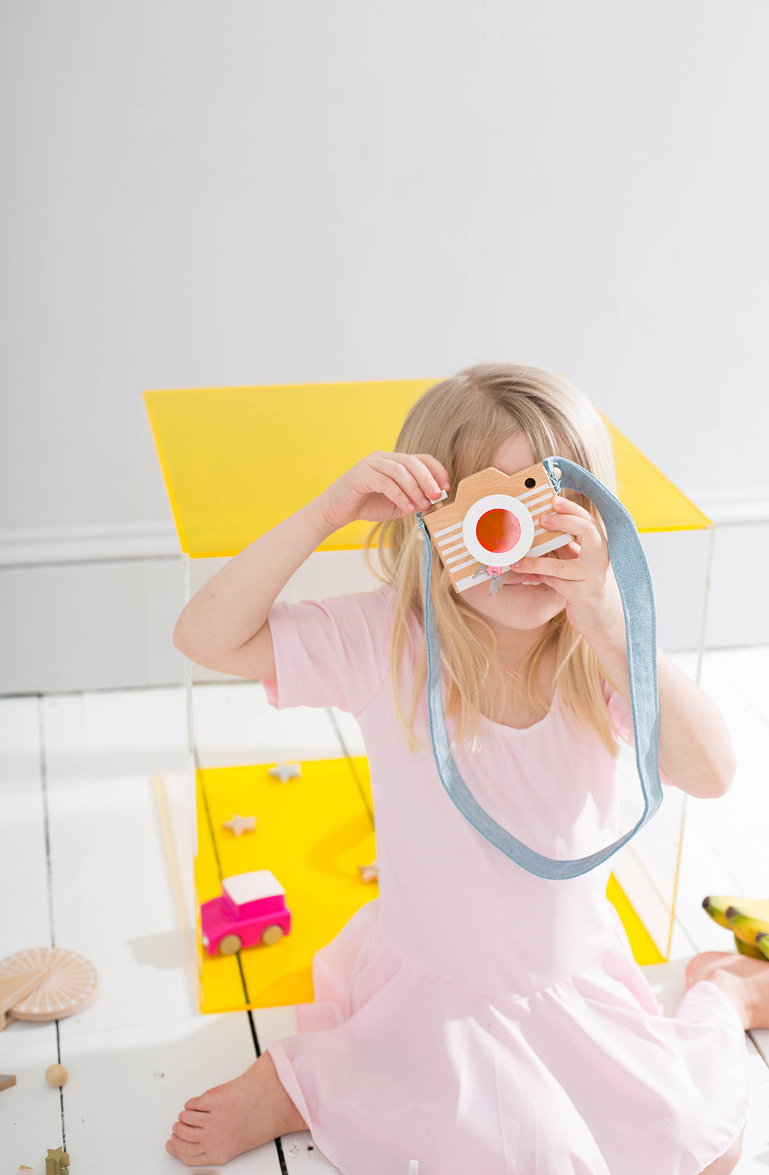 A young child with long blonde hair, wearing a light pink dress, sits on a white floor looking through the Kaleidoscope Play Camera Pink by KIKO & GG, which features a blue strap. In the background, there's a bright yellow cube and toys scattered around, creating the perfect scene for an amateur toy photographer's playful shoot.