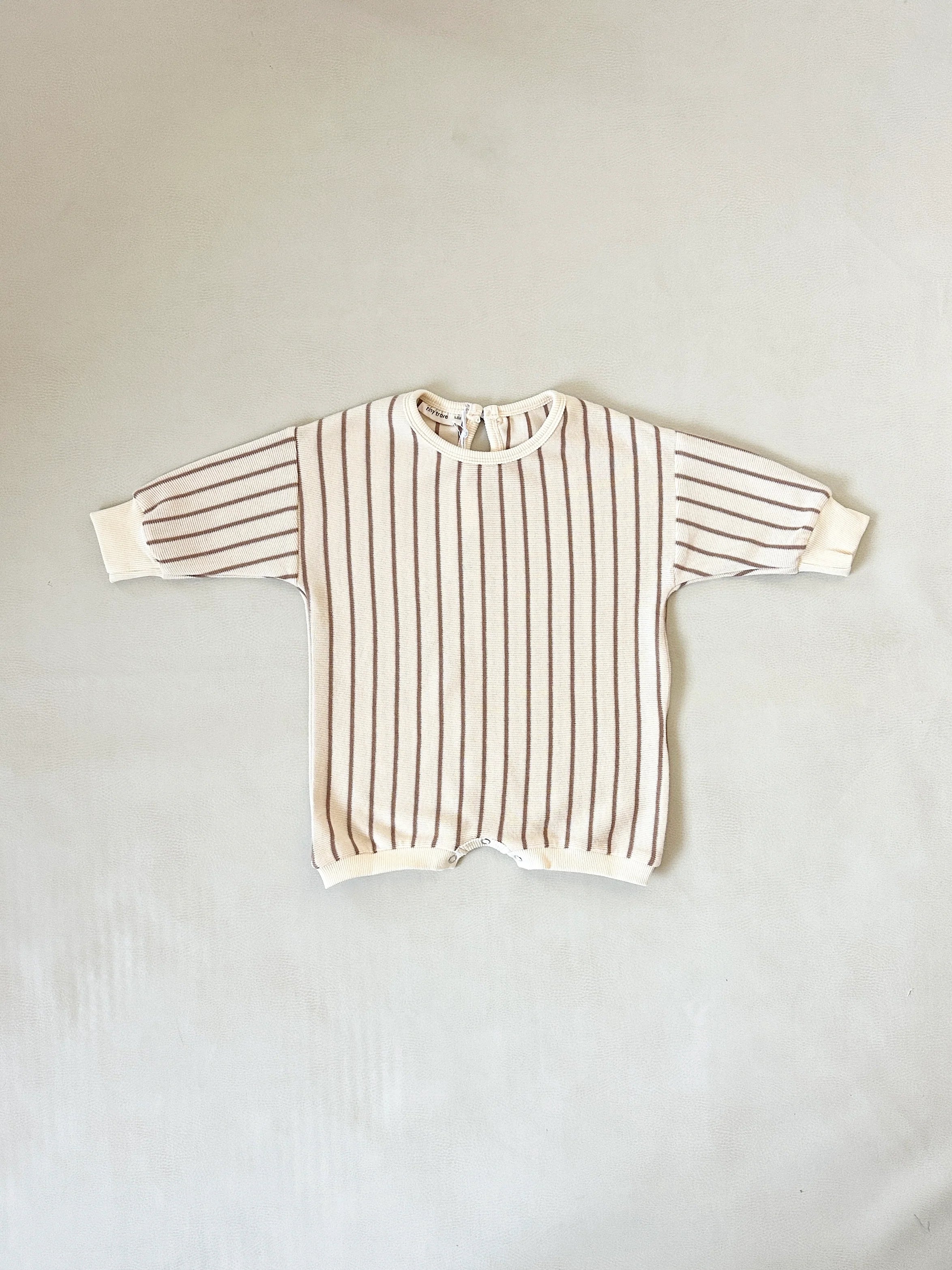 The TINY TROVE Tiny Trove - Addie Cotton Stripe Romper Cream/Latte Stripe is a cream-colored, long-sleeve 100% cotton romper with brown stripes, white cuffs, and a back neckline button, shown laid flat on a light background.