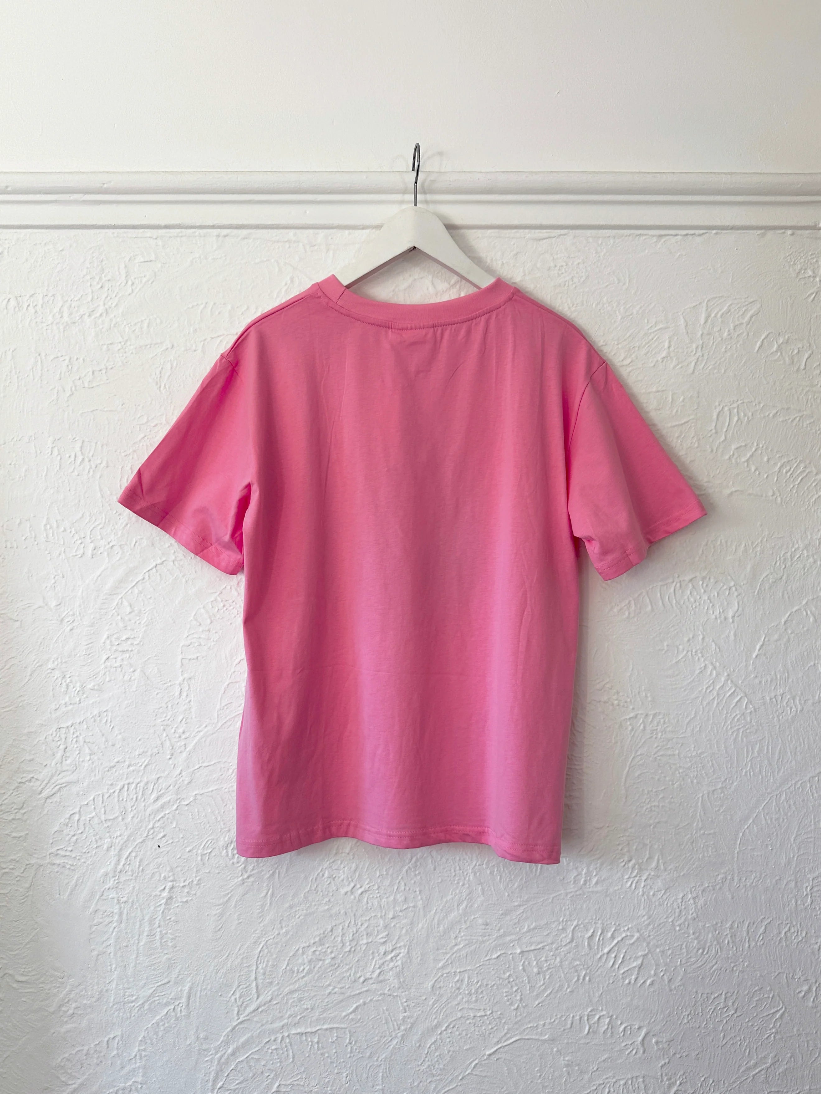 The Tiny Trove ~ Adult Farmer's Market Relaxed Tee Bubblegum by TINY TROVE, a plain pink cotton t-shirt with a relaxed fit, hangs backward on a white hanger. The shirt’s short sleeves and simple design stand out against the textured white wall with decorative patterns complementing the Tiny Trove print.