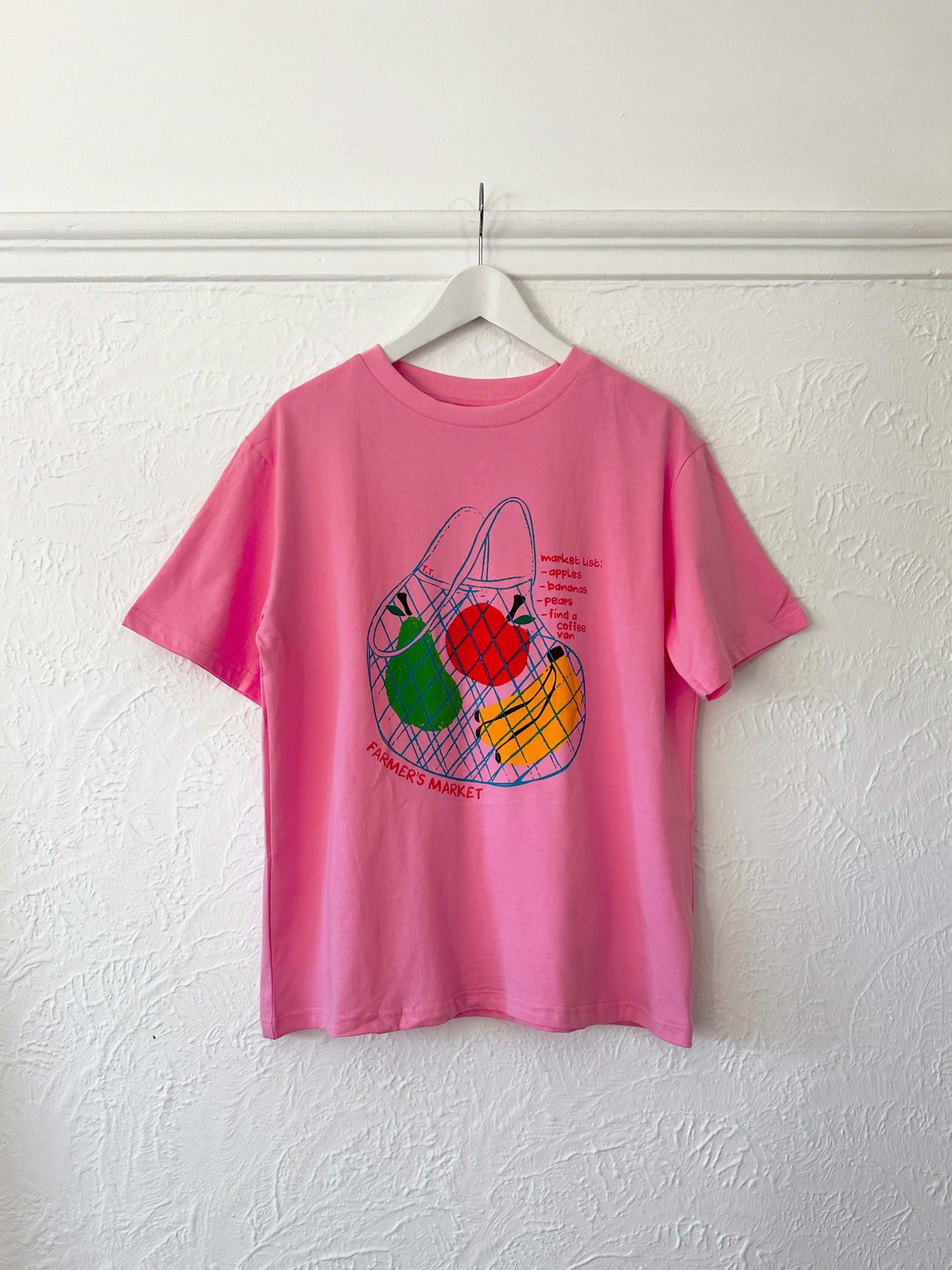 The Tiny Trove ~ Adult Farmer's Market Relaxed Tee Bubblegum, by TINY TROVE, hangs against a textured white wall. It showcases a net bag filled with colorful fruit and "Farmer's Market" text on a soft cotton pink T-shirt for ultimate comfort.