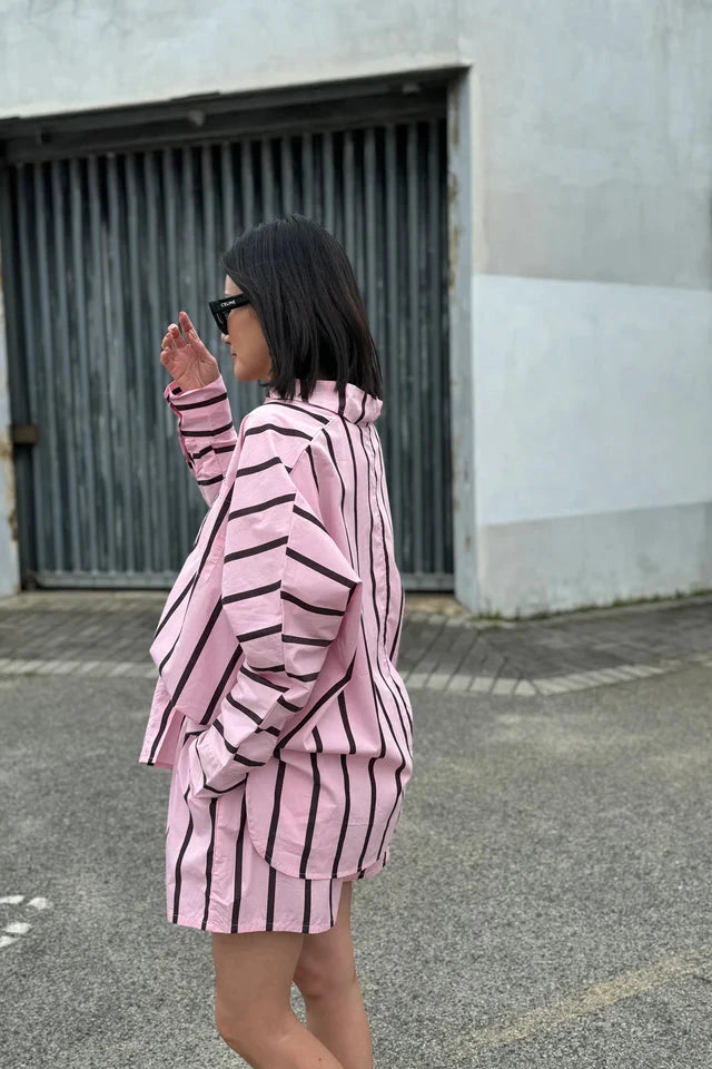 Someone with shoulder-length hair is wearing the Isla Stripe Cotton Set in Pink/Chocolate Stripe from TINY TROVE. The outfit, made of soft cotton, includes a long-sleeved shirt and shorts. They are standing outdoors in front of a large, closed garage door and holding their hand near their face.