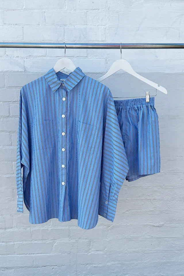 The Isla Stripe Cotton Set from TINY TROVE, featuring a relaxed-fit, button-up long-sleeve shirt and matching shorts in blue, white, and navy stripes, hangs on hangers against a white brick wall.