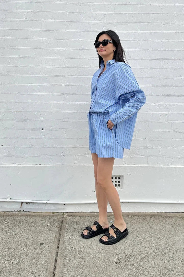 A woman wearing the Isla Stripe Cotton Set in Blue/White/Navy Stripe by TINY TROVE stands on a sidewalk in front of a white brick wall, sporting black sandals and sunglasses with her hands casually tucked into her pockets.