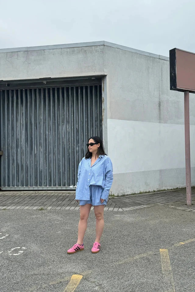 A person stands in a parking lot wearing the Isla Stripe Cotton Set by TINY TROVE, which features a relaxed fit and loose blue, white, and navy stripes. They pair this with pink shoes and sunglasses, posing confidently in front of a building with a large metal door and an empty billboard beneath an overcast sky.