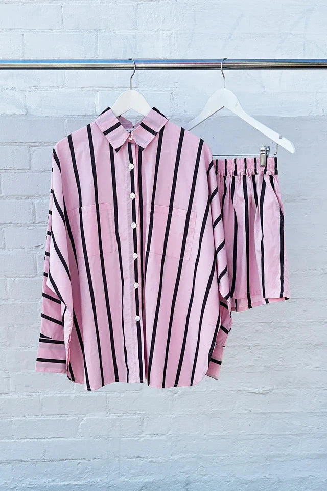 The Isla Stripe Cotton Set Pink/Chocolate Stripe from TINY TROVE, composed of a button-up shirt with black vertical stripes and crafted from breathable cotton, hangs on a metal rack next to the matching shorts. Both pieces offer a relaxed fit and are machine washable, set against a white brick wall.