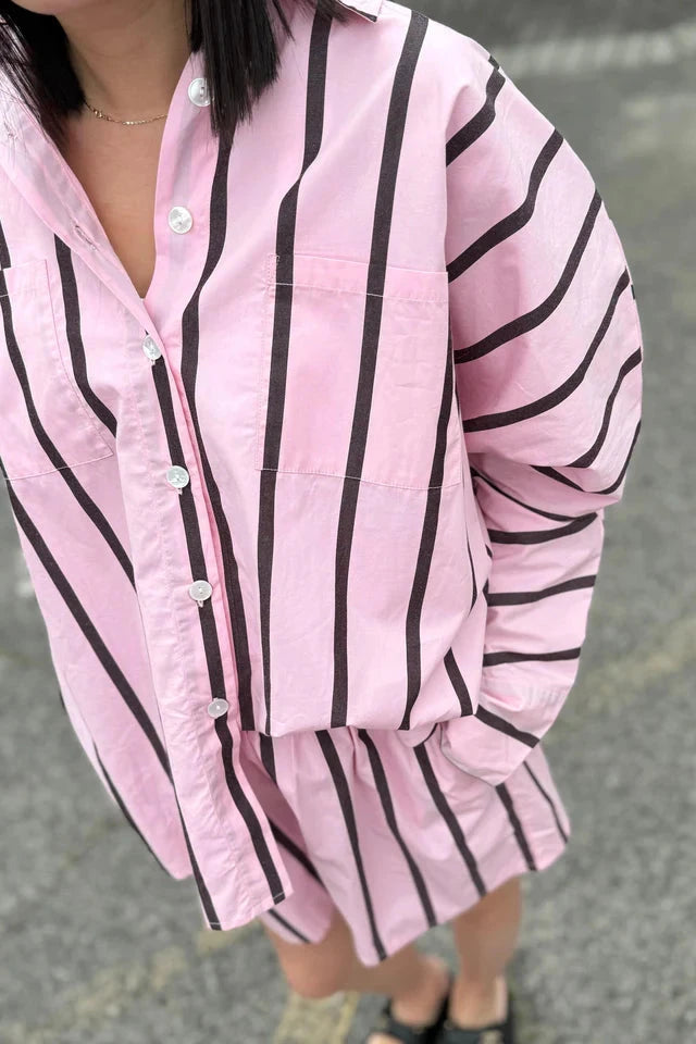 Dressed in the Isla Stripe Cotton Set from TINY TROVE, featuring a relaxed fit pink shirt with chocolate vertical stripes and matching shorts, a person stands against a gray textured background. The buttoned long-sleeve top pairs effortlessly with open-toed sandals, while one hand rests casually in the shorts pocket. The set is machine washable for easy care.