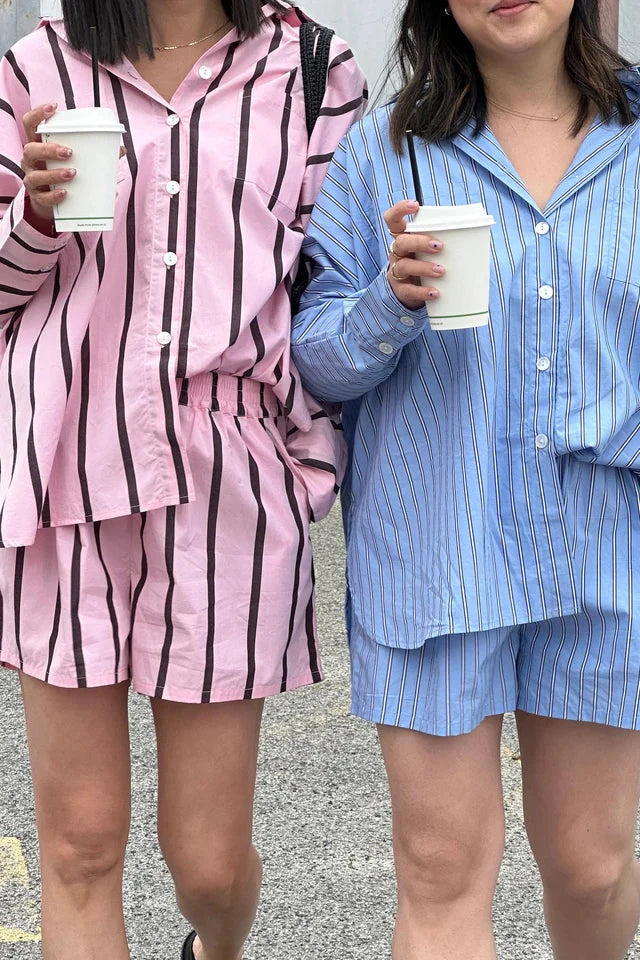 Two individuals wearing matching relaxed-fit outfits from TINY TROVE hold white coffee cups. One is dressed in the Isla Stripe Cotton Set featuring a blue, white, and navy stripe pattern, while the other wears a similar set in pink. They stand on a paved surface, each with one hand in their pocket.