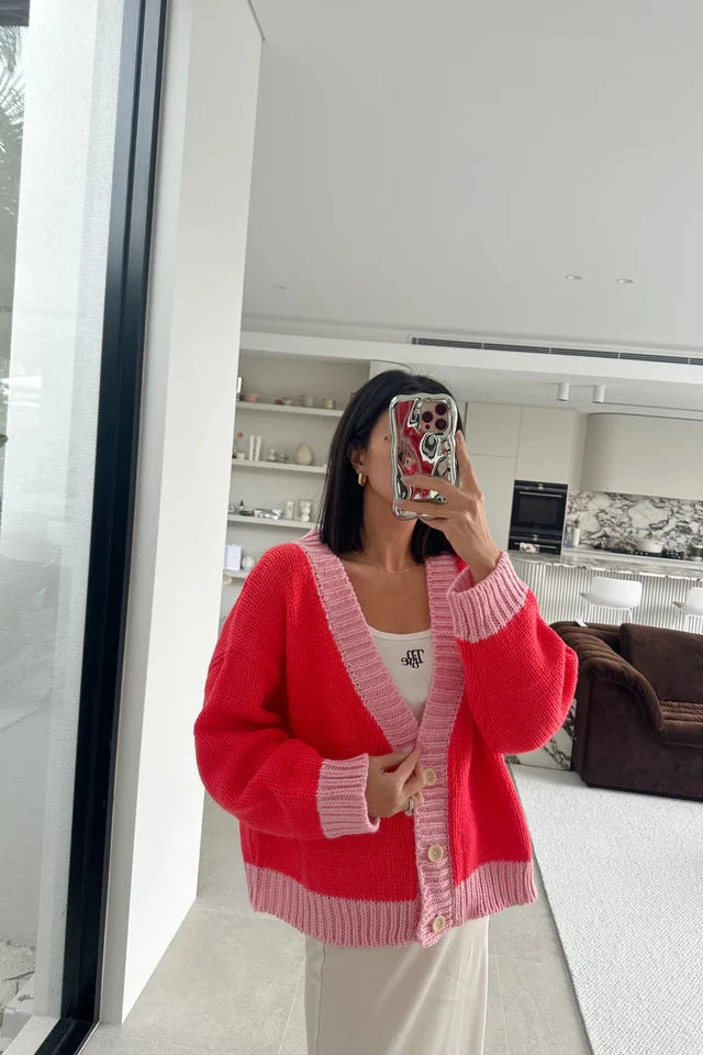 A person is taking a mirror selfie in a modern living room, sporting an oversized fit with the Adult Maeve Contrast Knit Cardigan in Red/Pink from TINY TROVE over a white top and beige pants. Their phone case features a red and white character design. The room has white walls, minimalistic decor, and a brown couch.