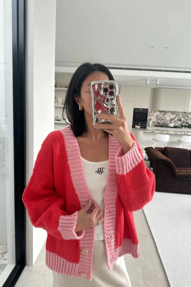 A person with shoulder-length hair is taking a mirror selfie with a phone that has a reflective case. They are wearing the Adult Maeve Contrast Knit Cardigan in Red/Pink from TINY TROVE over a white top with small black letters on the chest. In the background, there is a modern, neutral-toned living area.