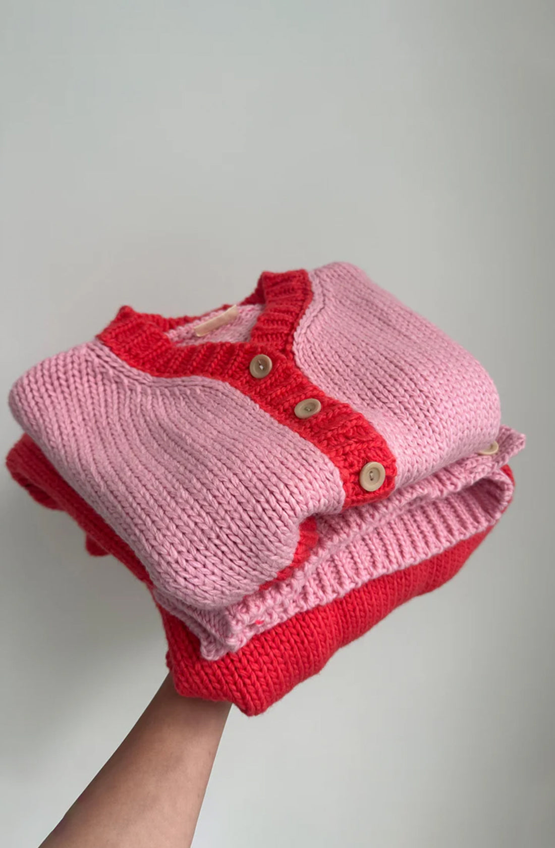 A hand holds a neatly folded stack of chunky knit sweaters. The top sweater, the Zuri Contrast Knit Cardigan Pink/Red from TINY TROVE, boasts an oversized fit with its pink body, red trim, and wooden buttons. The bottom sweater is red. Both are crafted from recycled acrylic and set against a plain and out-of-focus background.