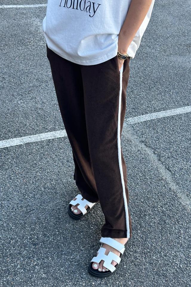 A person standing on a paved surface, wearing a white t-shirt with "Holiday" printed on it, styled with Piper Racer Pants in Dark Chocolate from TINY TROVE. The pants feature white side stripes and an elasticated waistband for comfort. Their relaxed leg style adds to the casual look, paired perfectly with white sandals. The focus remains on their legs and laid-back attire.