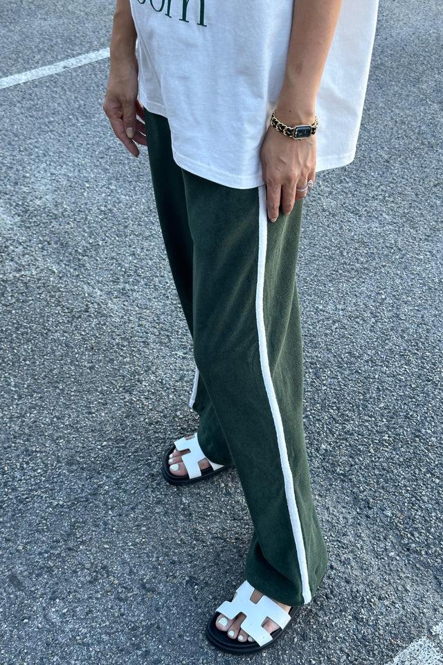 A person is wearing a white shirt, Piper Racer Pants Moss by TINY TROVE featuring white side stripes and an elasticated waistband, along with white sandals showcasing an interlocking "H" design. They are standing on a pavement, with their left arm adorned with a bracelet. Only the lower half of their body is visible in the image.