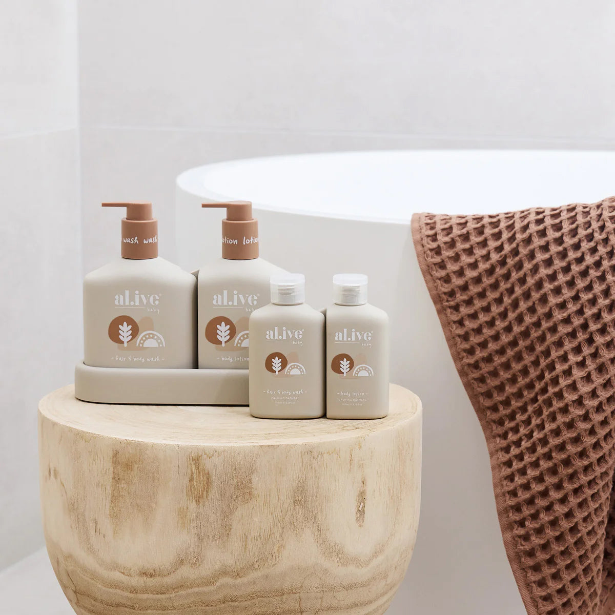 The al.ive body little traveller wash and lotion travel set infront of the standard sized body wash and lotion set. They are all sitting on a wooden table with a bath in the background with a waffle textured towel hung over the edge of the bath.