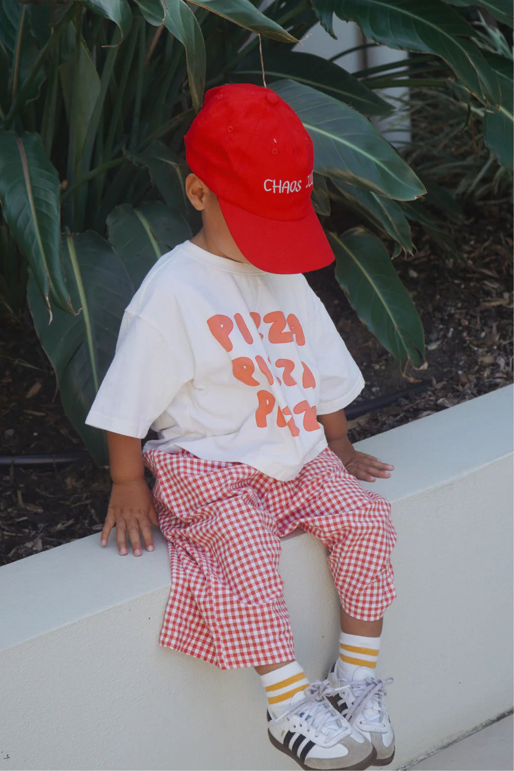 A young child sits on a low concrete wall in front of tropical leaves, wearing a white "Pizza" shirt, red cap, and WORN CURATIONS All Day Pant Chilli—unisex gingham pants featuring a comfy elastic waistband.