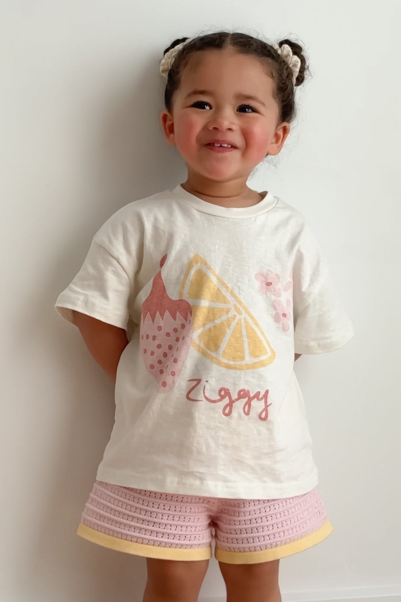 A young child with braided hair smiles while standing against a light background, wearing a white Print Tee August from ZIGGY LOU, adorned with fruit illustrations. Their pink shorts add a playful touch to the outfit, making it perfect for the Summer Collections.