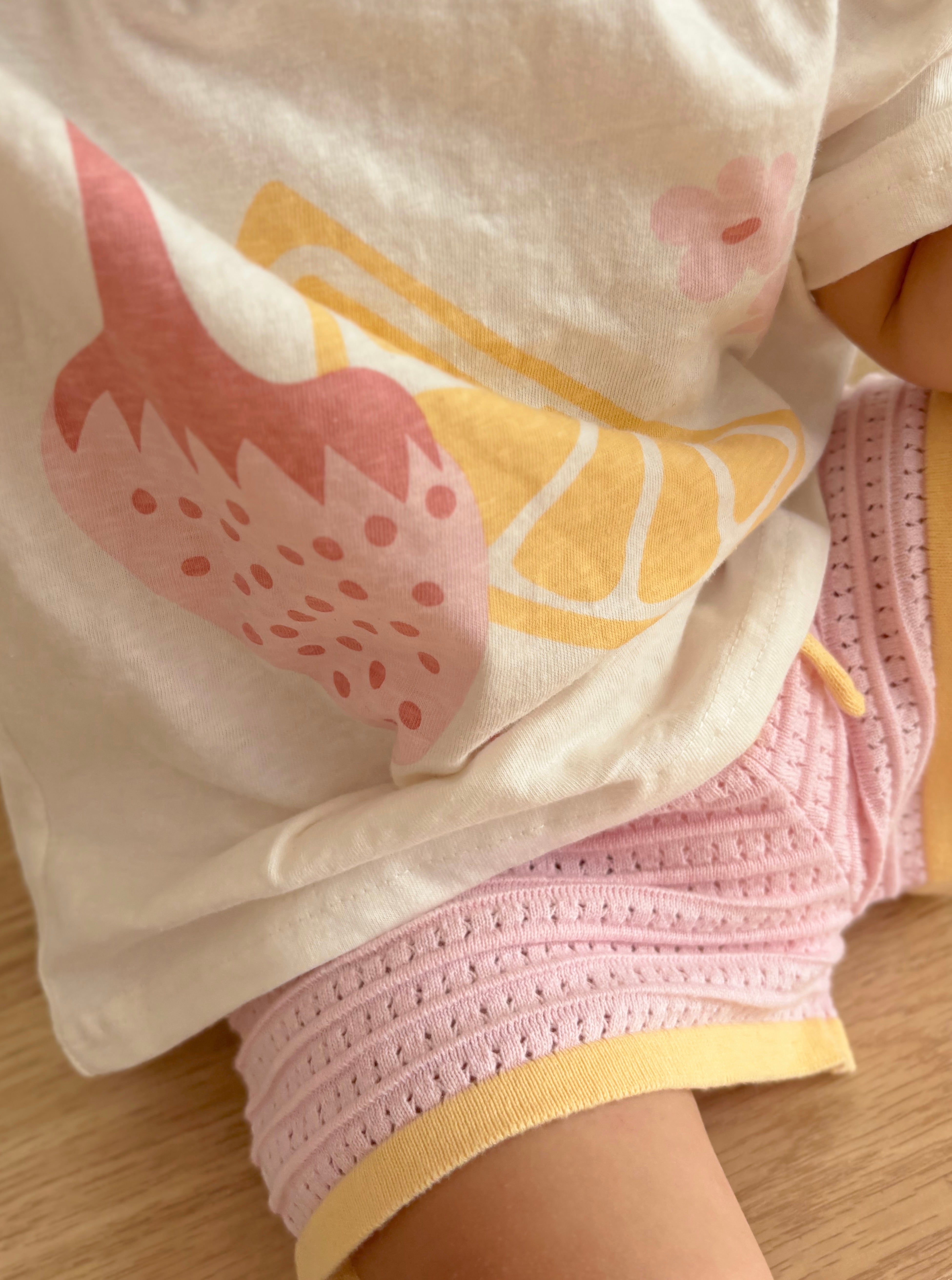 A child is dressed in a "Print Tee August" from ZIGGY LOU, showcasing a white shirt adorned with an August print of pink and yellow fruits. This is paired with pink shorts accented by yellow trim, all part of the Summer Collections. The outfit features an oversized fit as they sit comfortably on a wooden surface.