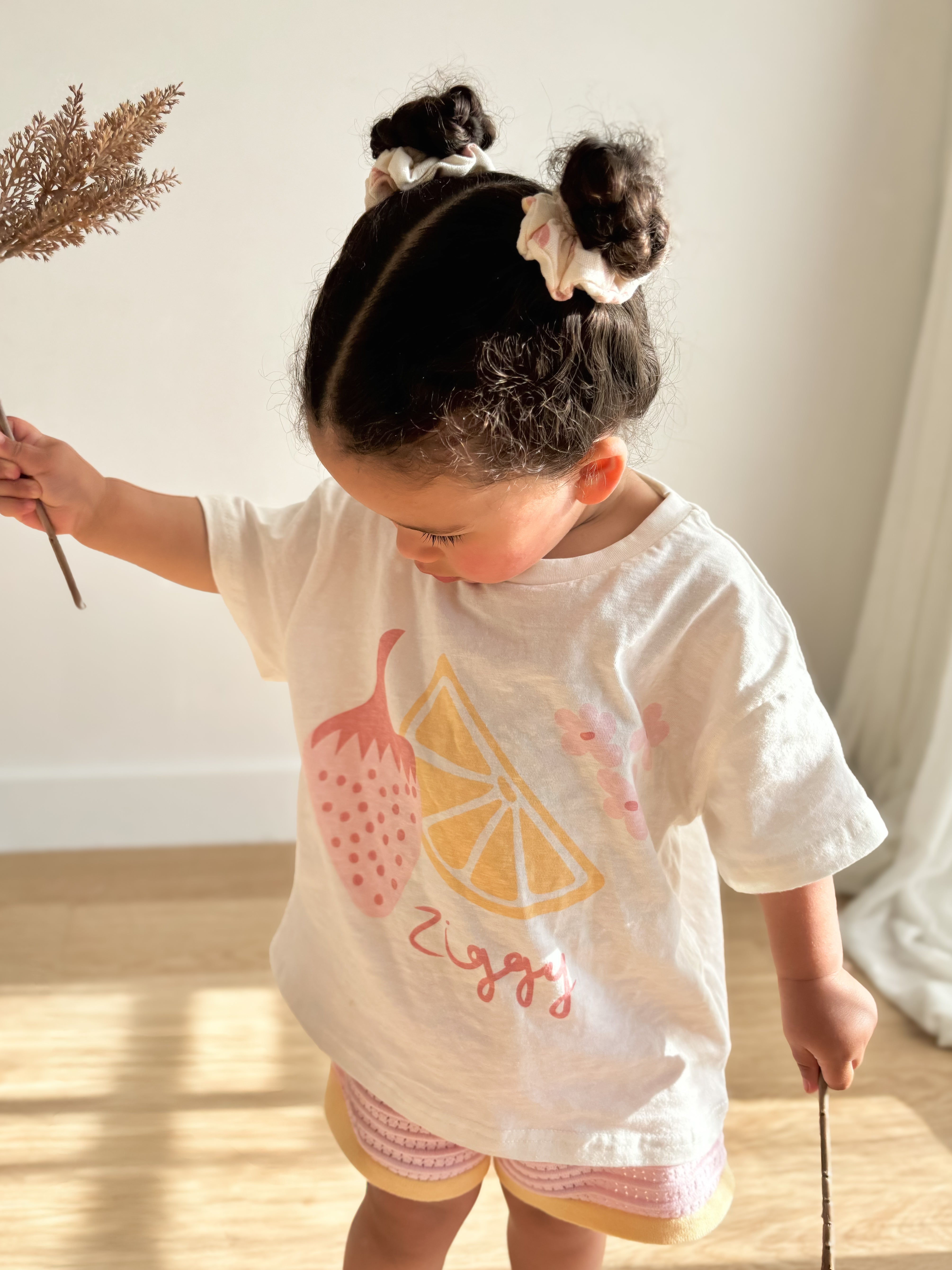 A young child stands indoors, dressed in an oversized fit white t-shirt from the ZIGGY LOU Summer Collection featuring fruit illustrations and the name "Ziggy" prominently displayed. They hold a brown twig in each hand, with their hair styled into two buns using light-colored scrunchies. The room is softly illuminated by natural lighting.