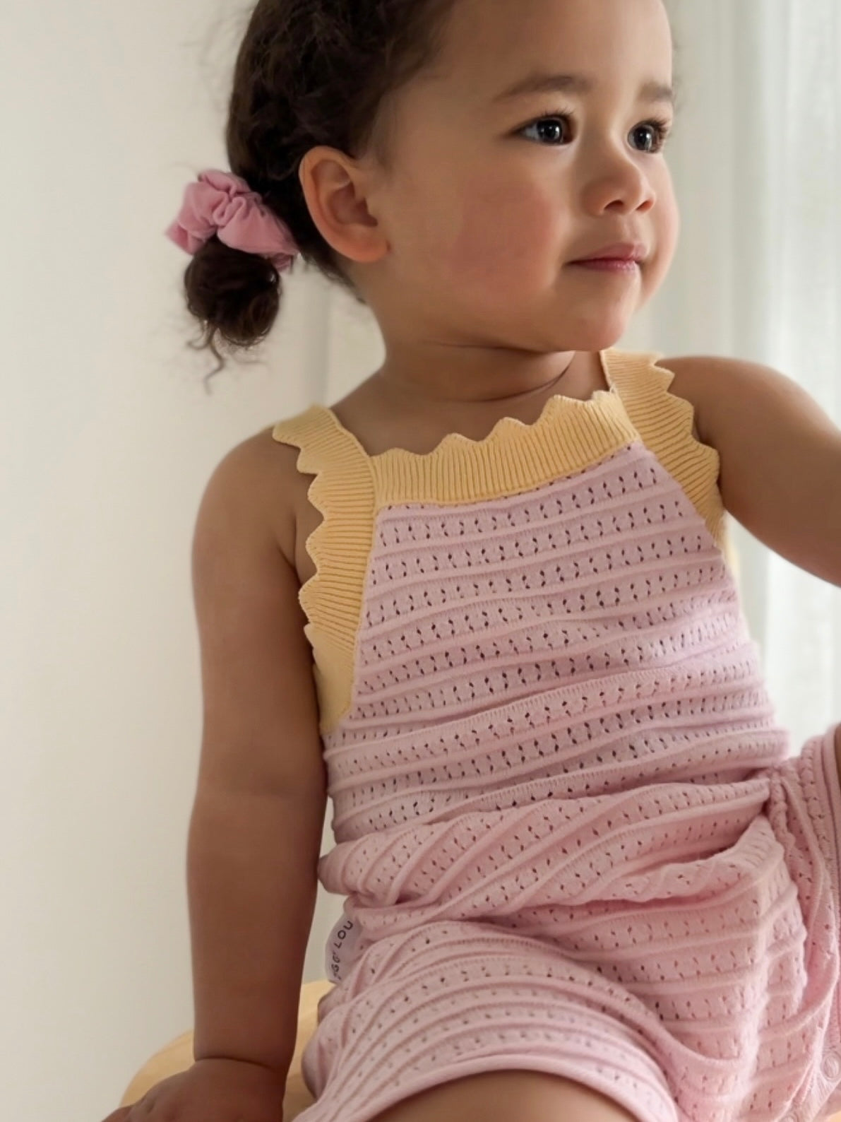 A toddler with curly hair tied in a pink scrunchie sits wearing the Playsuit Clementine by ZIGGY LOU, crafted from 100% cotton. The pink and yellow sleeveless romper perfectly complements her smile as she gazes to the side against a softly lit background, embodying the essence of cozy baby clothing.