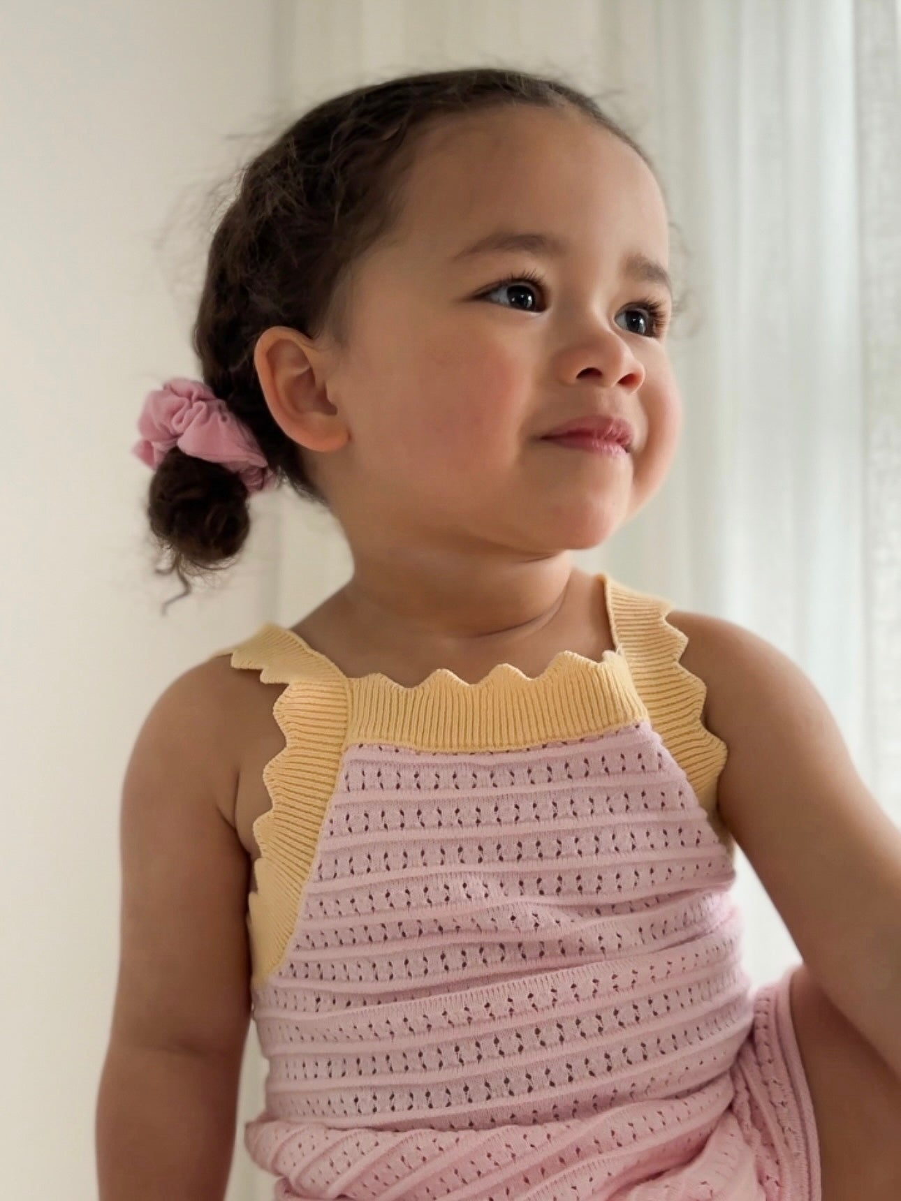 A young girl with curly hair in a ponytail, secured with a pink scrunchie, is wearing the charming Playsuit Clementine by ZIGGY LOU, which showcases yellow ruffled straps. She smiles softly as she looks to the side against a white curtain backdrop.