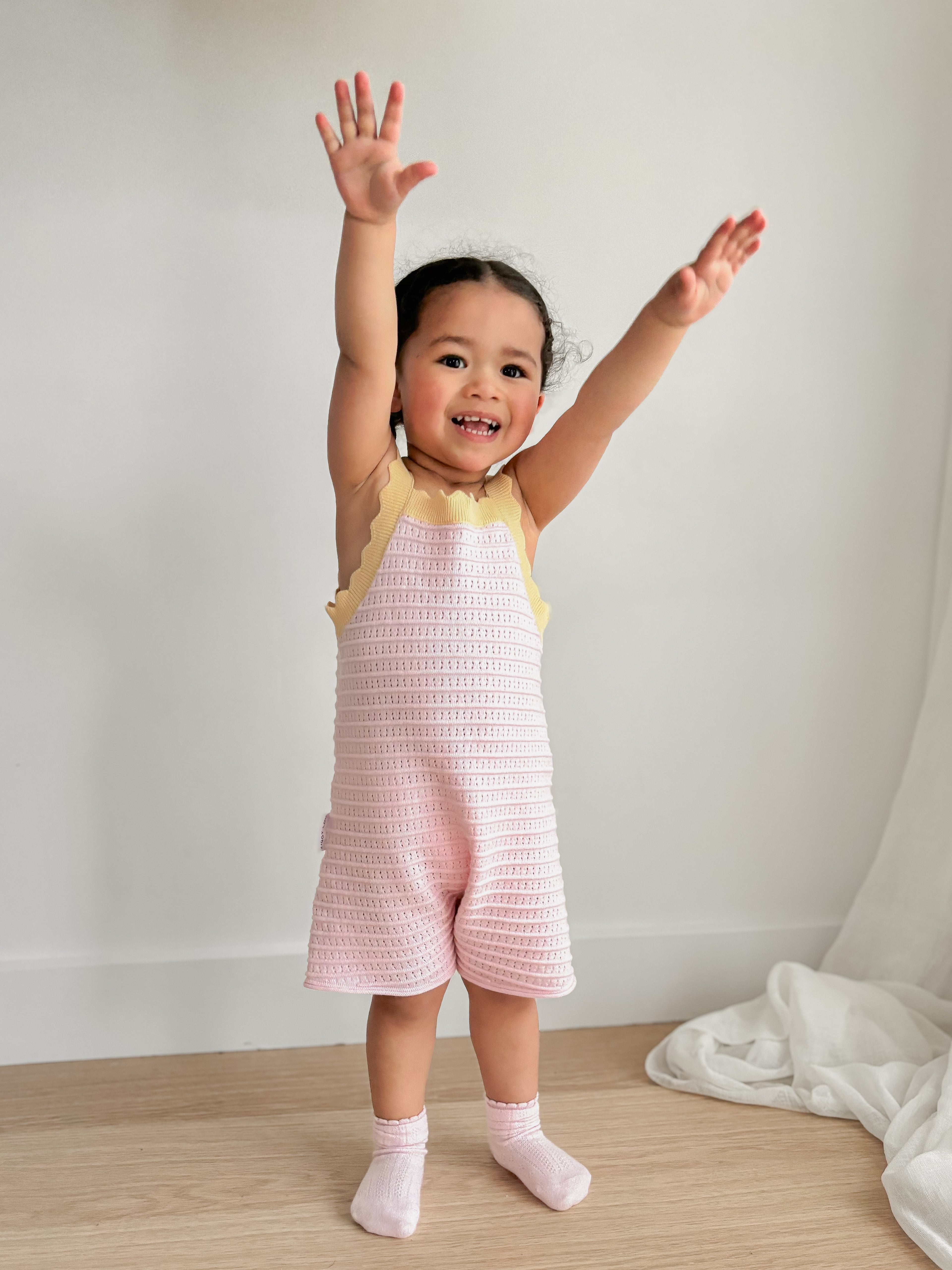 A young child with curly hair stands smiling, arms raised in the air. They are styled in the adorable ZIGGY LOU Playsuit Clementine, an eyelet knit design crafted from 100% cotton, layered over a pale yellow top. With pink socks completing the look, they add cheer to the minimalistic white room featuring wooden flooring and a draped white cloth backdrop.