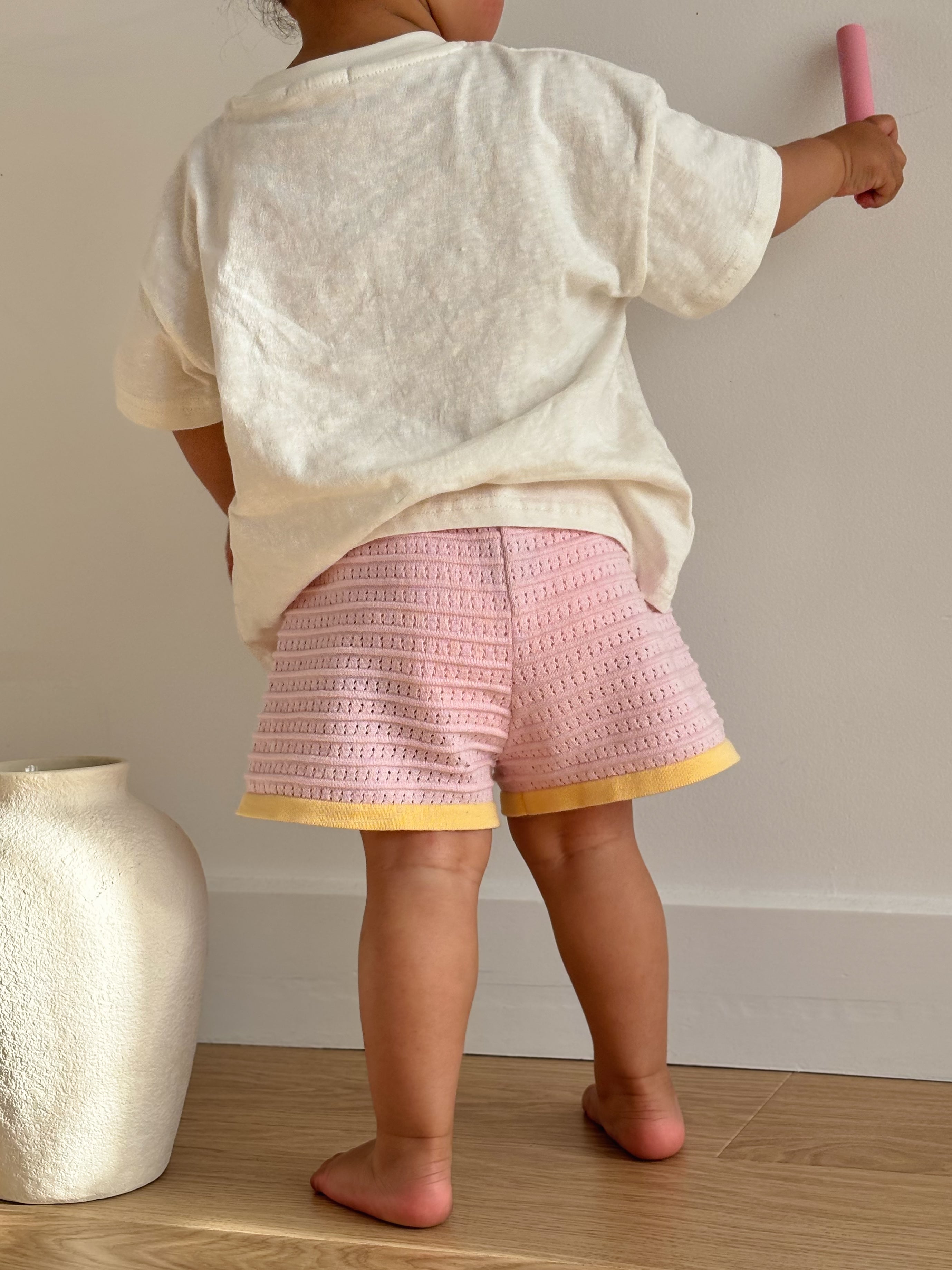 A child wearing a white T-shirt and Clementine shorts from the ZIGGY LOU Summer Collection stands on a wooden floor. They hold a pink chalk stick against a white wall, with a white textured vase partially visible on the left.