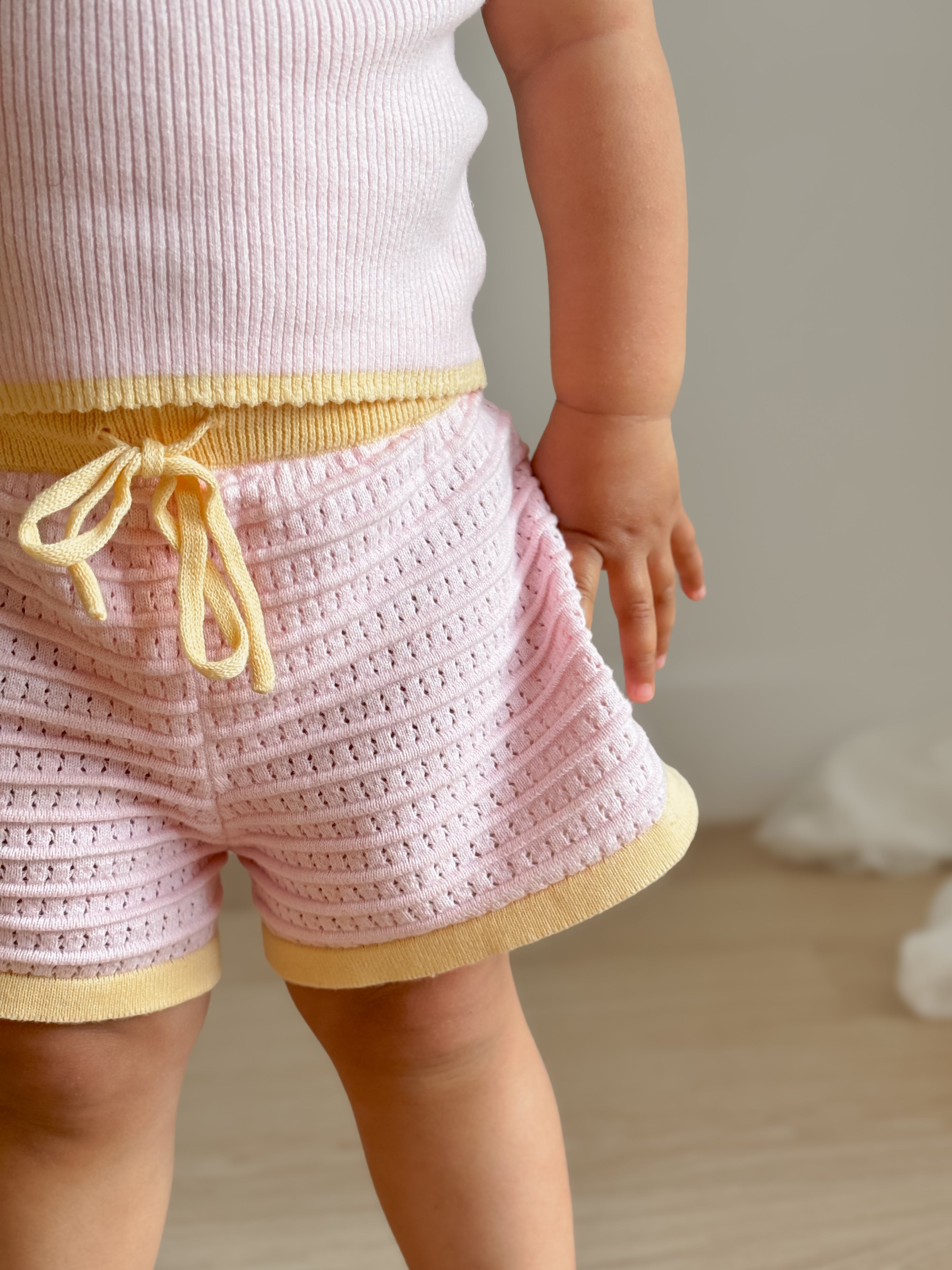 A toddler dressed in a pink knitted tank top and the Shorts Clementine by ZIGGY LOU, adorned with eyelet detail and yellow trim, stands on a wooden floor, perfectly capturing the charm of the Ziggy Lou Summer Collections. Their hand rests by their side as they enjoy the comfort and softness provided by 100% cotton.