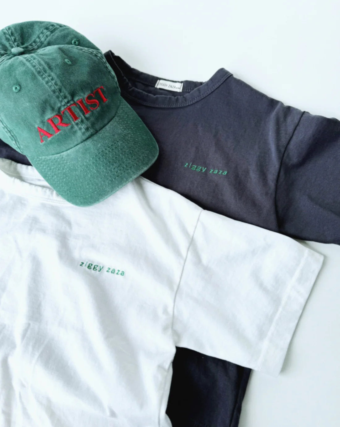 Two t-shirts, one white and one black, from ZIGGY ZAZA lay on a white background. The Ziggy Zaza ~ Artist Cap in Forest Green, featuring pre-washed cotton canvas with an adjustable brass clasp and "ARTIST" in red, is stylishly placed on the black shirt.