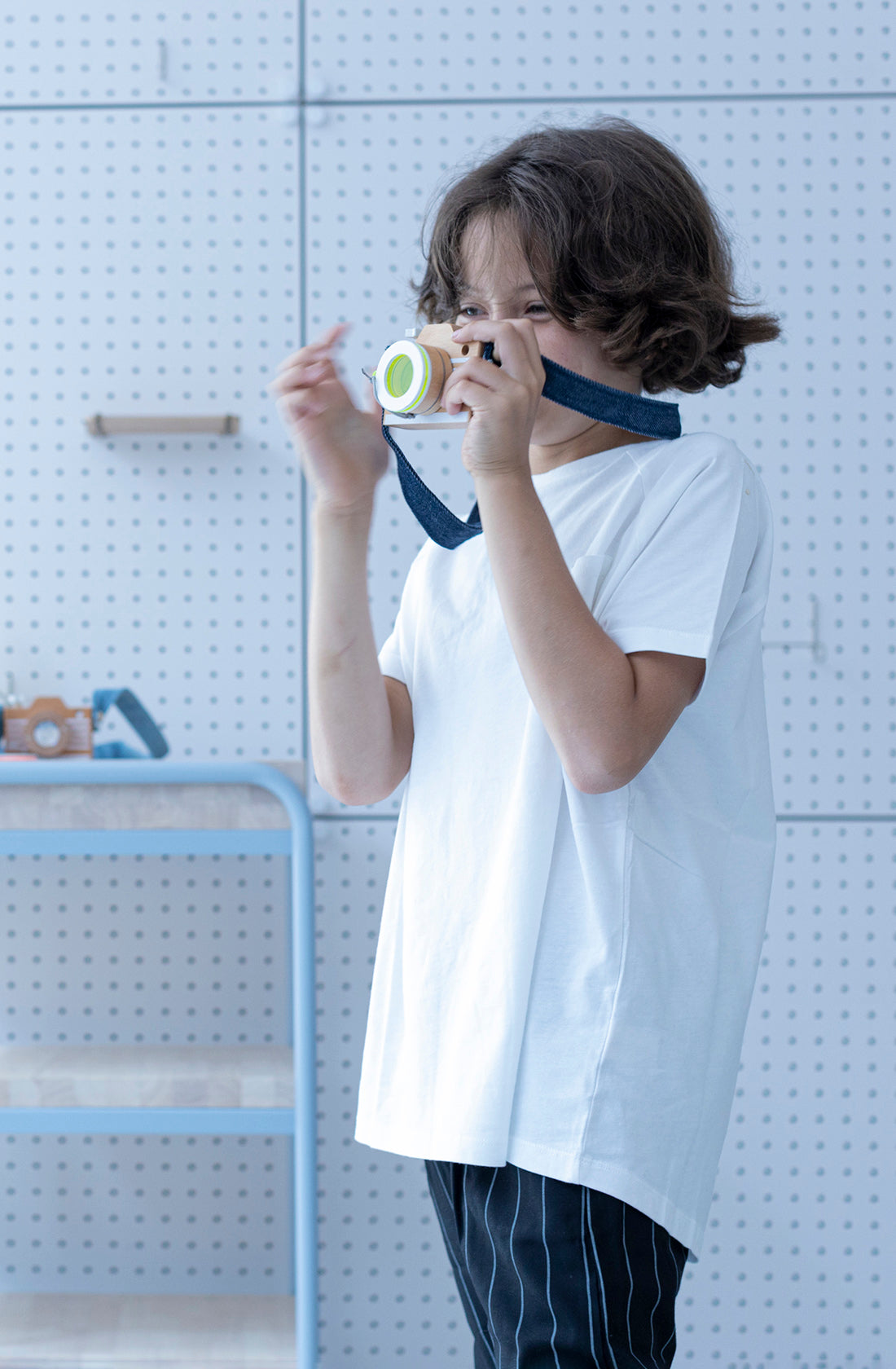 A young child with curly hair, dressed in a white t-shirt and dark striped pants, is holding the Kaleidoscope Play Camera Yellow by KIKO & GG to their eye as if taking a picture. The background features light-colored pegboard walls and shelves. It's as if the little photographer has captured an unexpected kaleidoscope surprise.