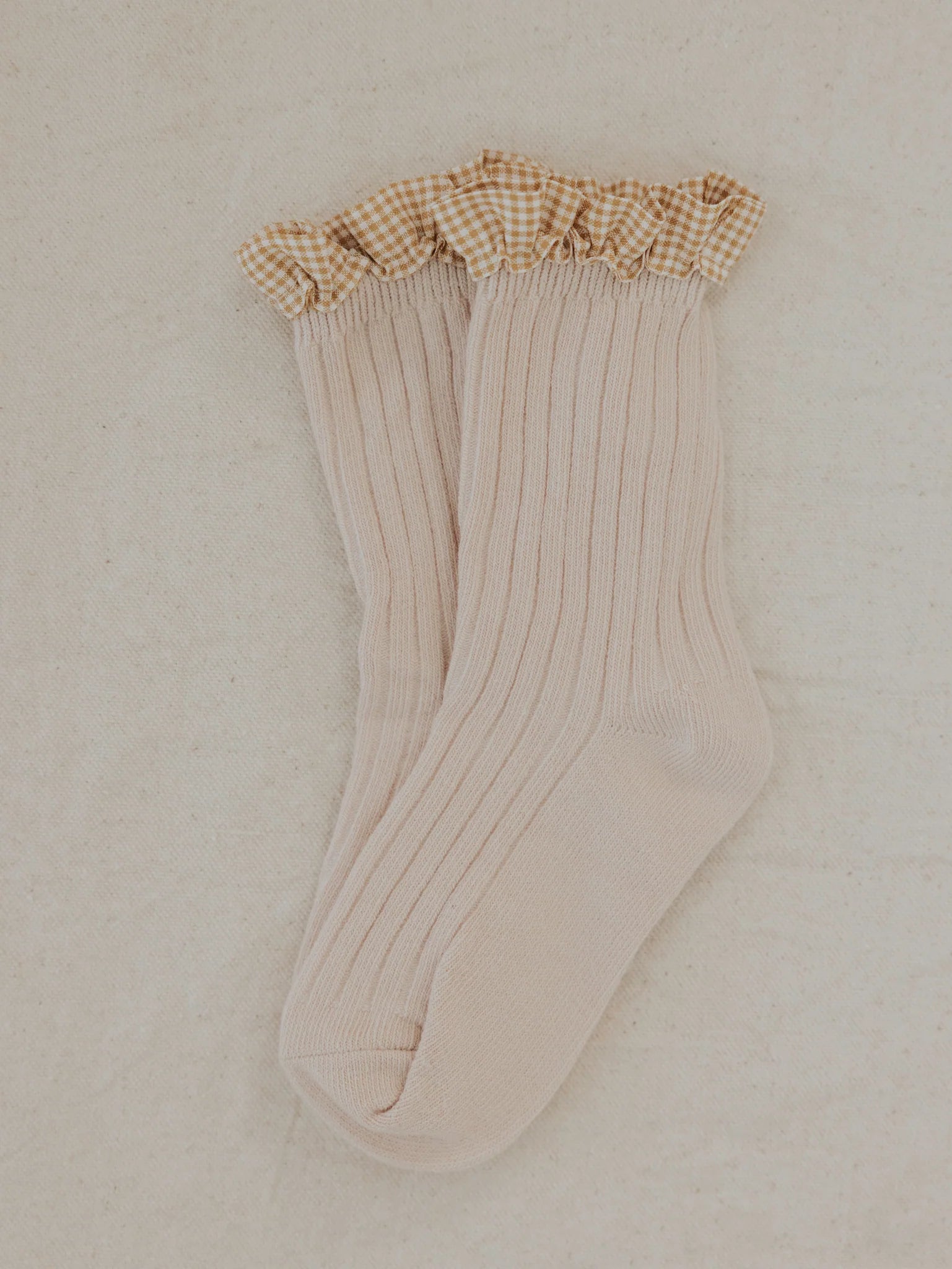 A pair of Frilly Socks Oat from VALENCIA BYRON BAY, featuring a light beige ribbed design and a charming brown-and-white checkered ruffled cuff, lies neatly side by side on a light-colored, textured fabric background. These cotton socks are perfect for little feet.