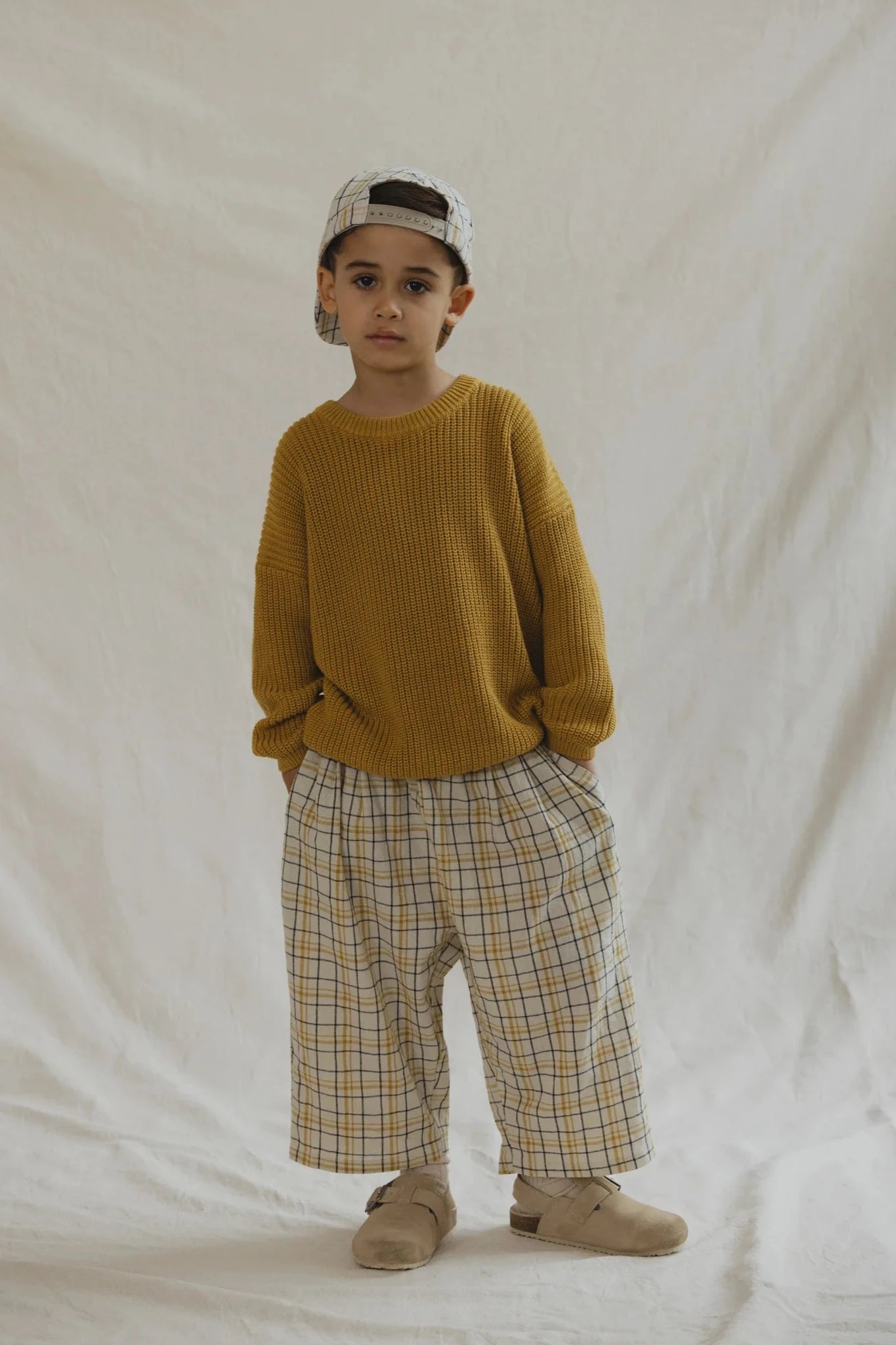 A young child stands against a plain backdrop, wearing an oversized mustard-yellow sweater, VALENCIA BYRON BAY's durable and practical Rio Pant Picnic plaid pants made from 100% cotton, beige shoes, and a backwards baseball cap. They have their hands in their pockets and a neutral expression.