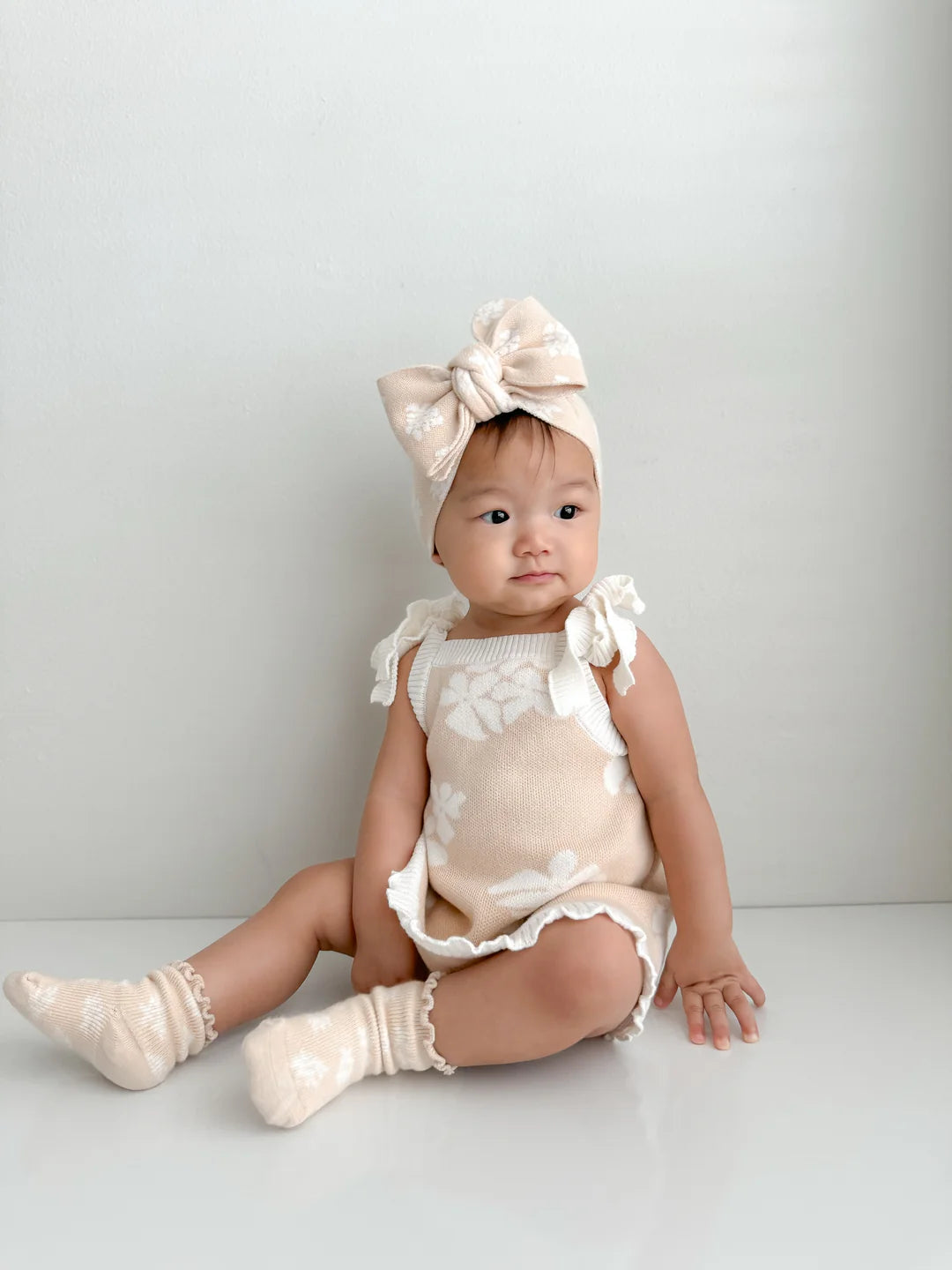 A young child wearing a beige outfit with white floral patterns sits against a plain background. The outfit, from the Banksy + Beech Spring Collections, includes a top with ruffled sleeves, matching Socks Banksy by ZIGGY LOU, and a large headband with a bow. The child is looking slightly to the side with a calm expression.