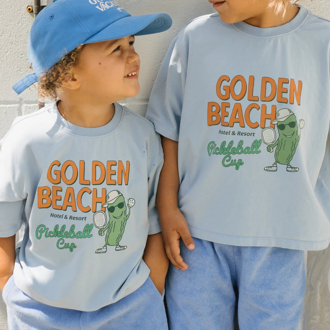 Two children sport relaxed fit Golden Beach Mid Sleeve Tees in Palm Blue by GOLDEN CHILDREN, each adorned with "Golden Beach Pickleball Cup" and a cartoon pickle playing pickleball. One child wears a blue cap as they both share smiles against a light backdrop.