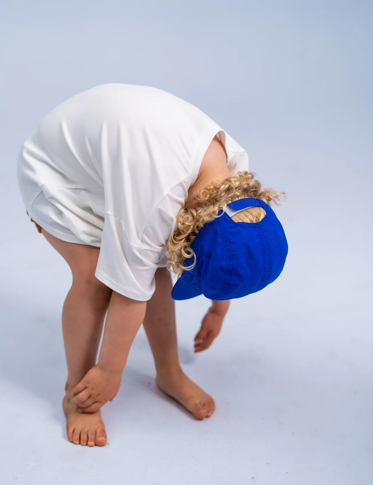 A child wearing the Summer Club Cap: Electric by SONNY LABEL, featuring an adjustable velcro strap, and dressed in a white t-shirt bends forward to touch their feet against a light blue background.