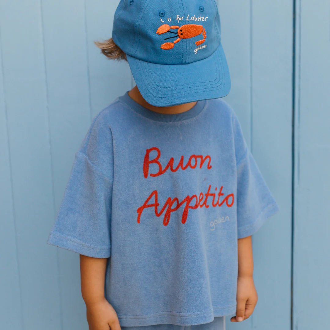 A child wearing a stylish Lobster Cap Deep Sea Blue from GOLDEN CHILDREN, featuring an adjustable brass clasp and the text "L is for Lobster," pairs it with a blue shirt displaying red "Buon Appetito" lettering. The background complements perfectly with its matching blue hue.