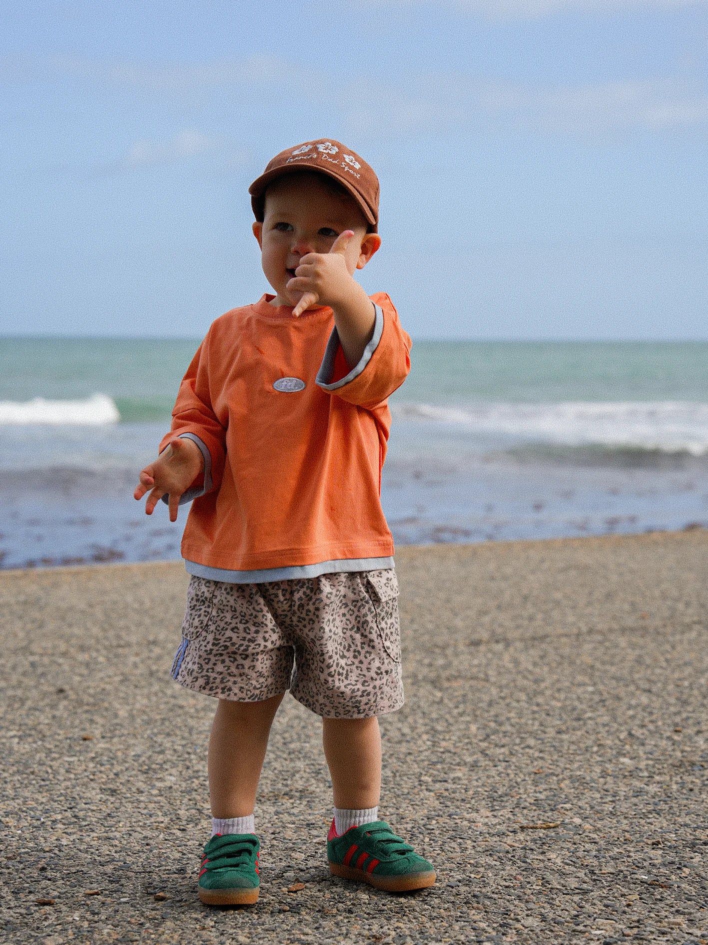 A toddler in a slightly large PRE-ORDER Double Layer Tee Paw Paw by FRANCO'S DAD, paired with patterned shorts and a brown cap, stands on a beach promenade. With the ocean and blue sky as a picturesque backdrop, the child gestures with one hand while holding a small object in the other.