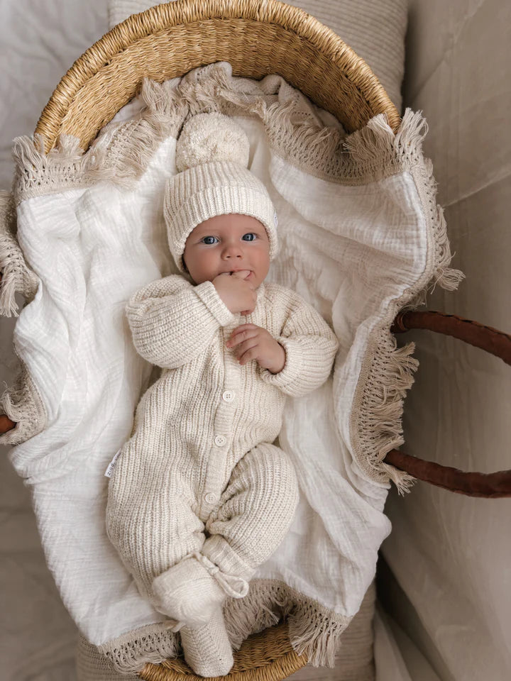 A baby lies in a woven bassinet wrapped in soft white blankets. Clad in the ZIGGY LOU Classic Knit Romper Honey, with a matching hat and mittens to complete the cozy cream-colored ensemble, the baby gazes up with bright eyes, gently sucking on their thumb. The scene exudes warmth and comfort, perfect for a winter outfit.