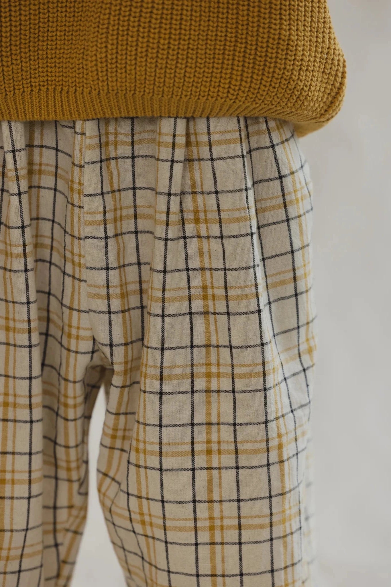 Close-up of a person wearing the VALENCIA BYRON BAY Rio Pant Picnic, crafted from 100% cotton with a yellow and black checkered pattern, paired with a mustard yellow knitted sweater. The image captures the texture and details of the garments, emphasizing the plaid design and knit fabric, showcasing durable and practical style.