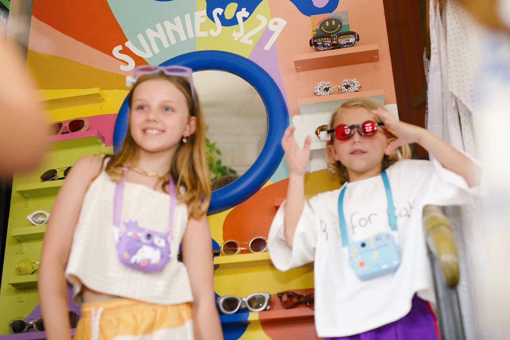 Two children stand before a vibrant sunglasses display, one in purple shades, the other in red, beneath a "SUNNIES $29" sign. Sporting small bags, they seem set for imaginative play or capturing memories with an Oh My Frankie ~ Oh My Snap Instant Print Camera Blue by OH MY FRANKIE.