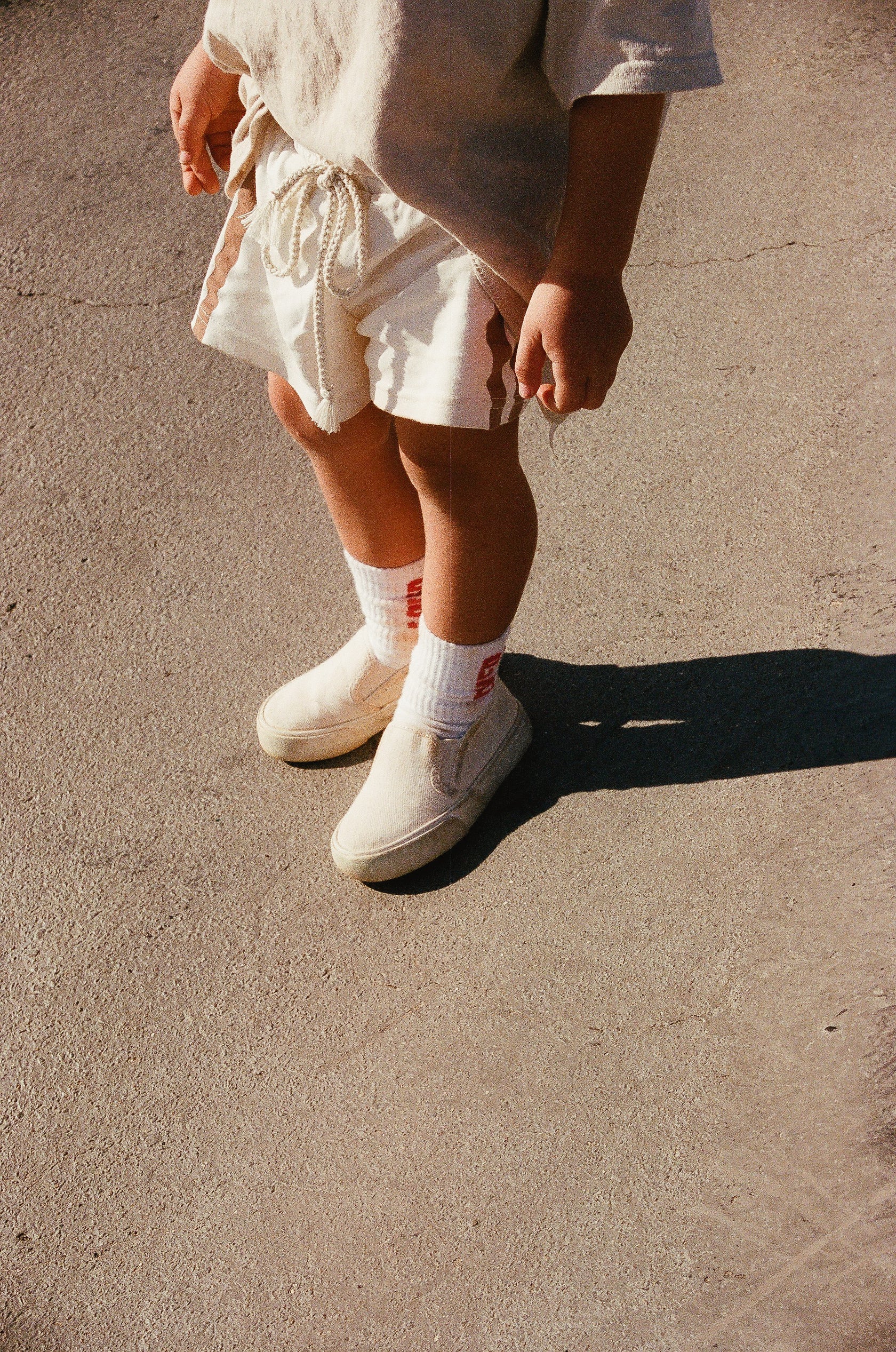 A child, aged 2-5 years, is standing on a concrete surface, dressed in beige shorts with a rope tie, a beige shirt, white TINY LOVE CLUB Loved Socks with red prints and elastic banded ankles, and beige slip-on shoes. The child is facing forward, and the image is taken from the shoulder down.