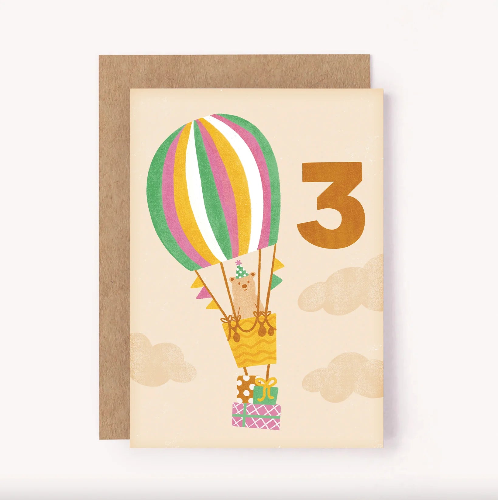 A Lauren Sissons Studio designed birthday card Featuring an illustrated bear in a striped hot air balloon, carrying a stack of three wrapped gifts. There is a bold hand-lettered number 3 on a soft beige background.