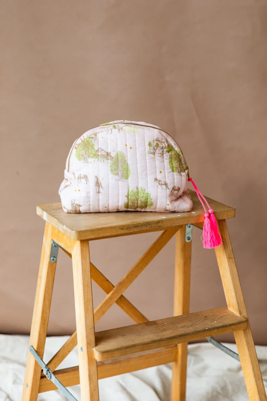 A half moon shaped purse with horses, trees and old houses on it with a pink tassle zip sitting on a wooden step ladder.