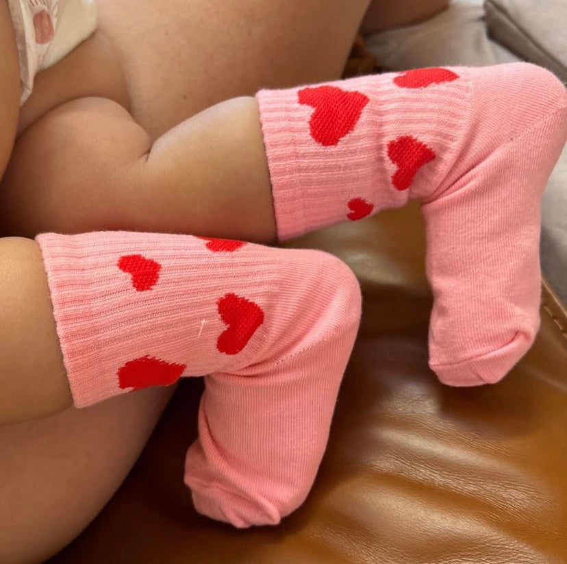 A baby wearing adorable TINY LOVE CLUB Burst Socks, decorated with red hearts, is lying down. The baby's legs are bent at the knees as they rest on a brown leather surface. The image highlights the baby's legs and the machine washable Burst Socks, with only part of the baby's body visible.