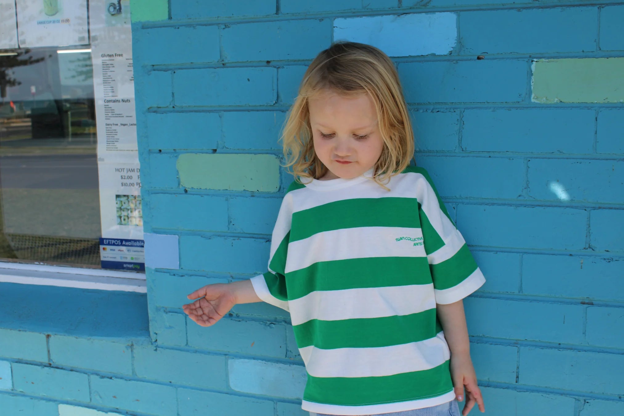 A young child with long blond hair stands against a blue-and-turquoise brick wall, wearing the Twin Collective's oversized green-and-white striped tee made from 100% cotton. The child gazes downward with one hand touching the wall, and there's a window to the left.