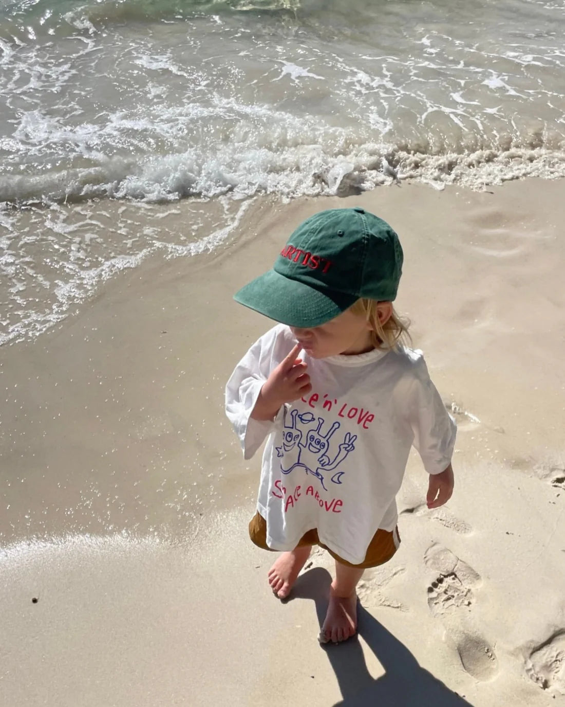 A child in a white shirt with colorful designs wears the Ziggy Zaza ~ Artist Cap Forest Green by ZIGGY ZAZA and stands barefoot on the sandy beach, gazing thoughtfully at the sand as gentle waves lap at the shore.