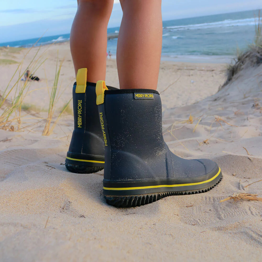 Close-up of a child's legs in "MERRY PEOPLE" Merry People ~ Andie Kids Gumboot Midnight Blue rubber boots, standing on sandy dunes with grass and ocean waves in the background. These vegan-friendly boots are perfect for beach adventures.