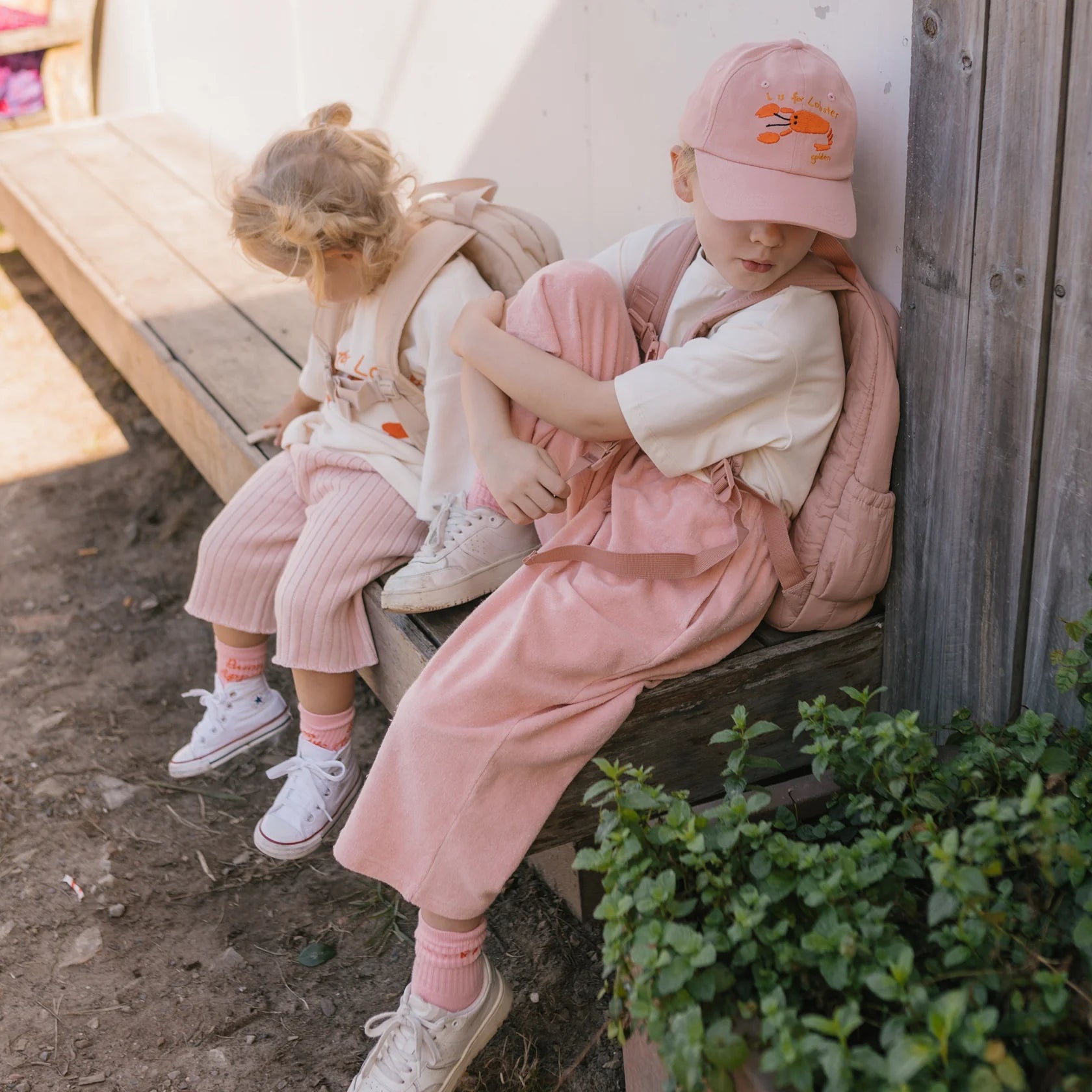 Two young children sit on a wooden bench outdoors. Both are dressed in pastel outfits, wearing GOLDEN CHILDREN Confetti Wide Leg Pants in the shade of Pink Parfait, white shirts, and matching pink sneakers. One child wears a pink cap and has a backpack, while the other has blonde hair tied back and also carries a backpack.